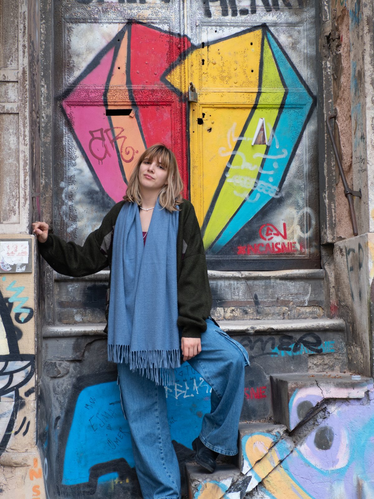 Leyla Fıratlı standing on graffiti-covered steps in front of a rainbow-painted door, wearing a blue scarf