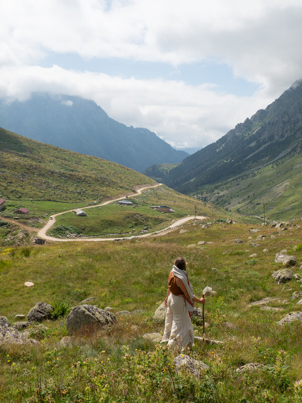Leyla standing in a high mountain meadow with a walking staff, looking down the valley at a winding dirt road