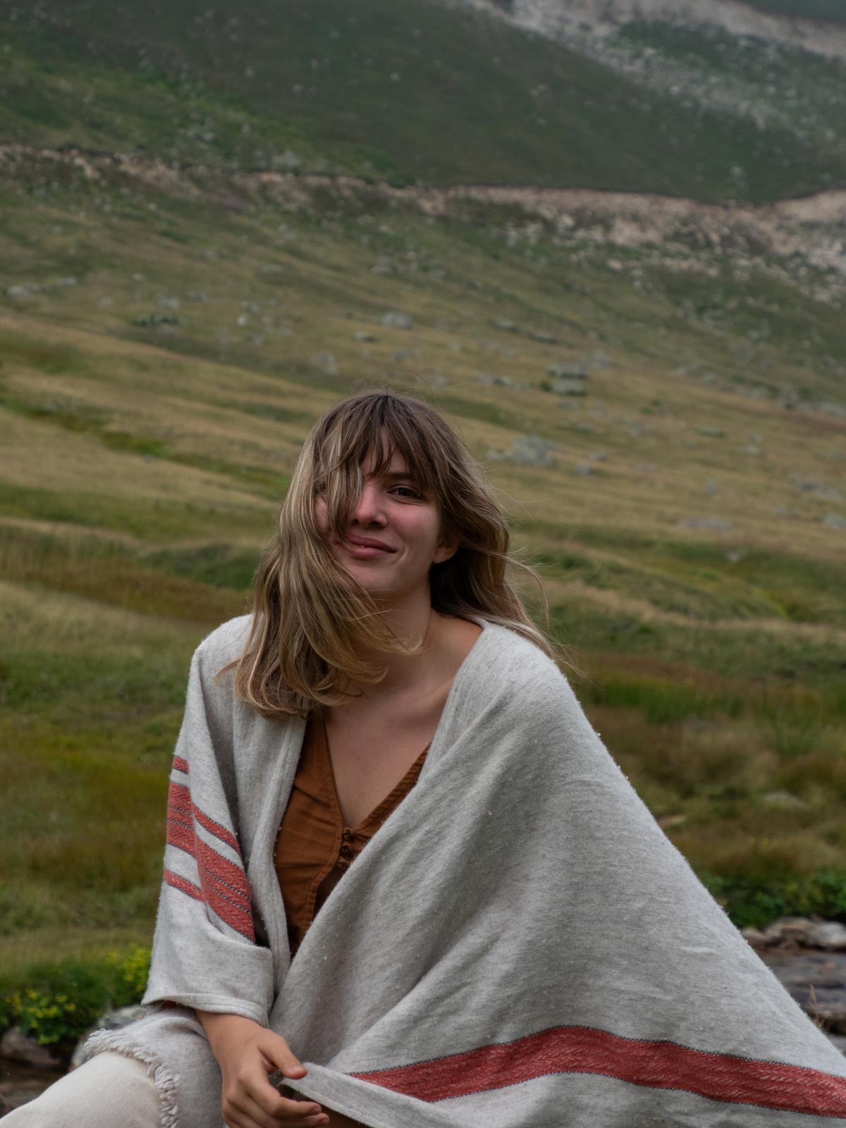 Leyla sitting on a green mountain plateau, wrapped in a cream and red blanket, smiling